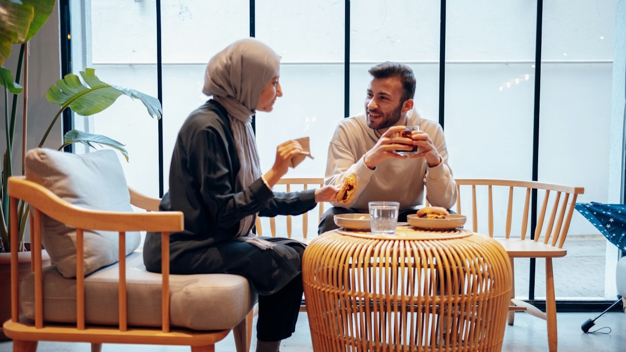 A woman wearing a hijab and a man are seated in a modern cafe, smiling at each other over coffee and a light meal on a round wicker table.