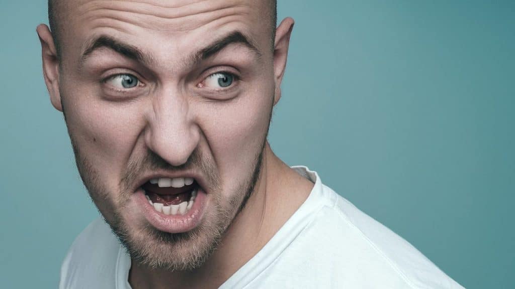 Close-up of a bald man with a beard looking angry and yelling on a blue background.