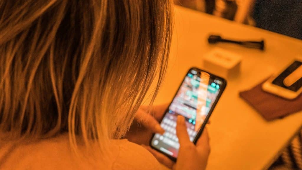 An over-the-shoulder shot of a person with blonde hair, looking down at and tapping on a smartphone screen on a brightly lit table.