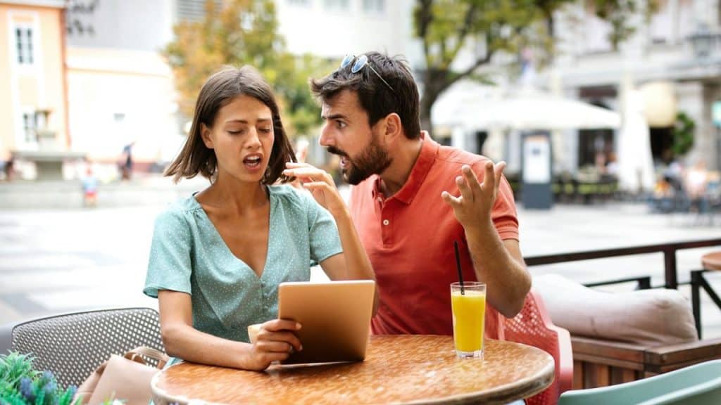 A woman looks annoyed holding a tablet while a man argues and gestures at an outdoor table.