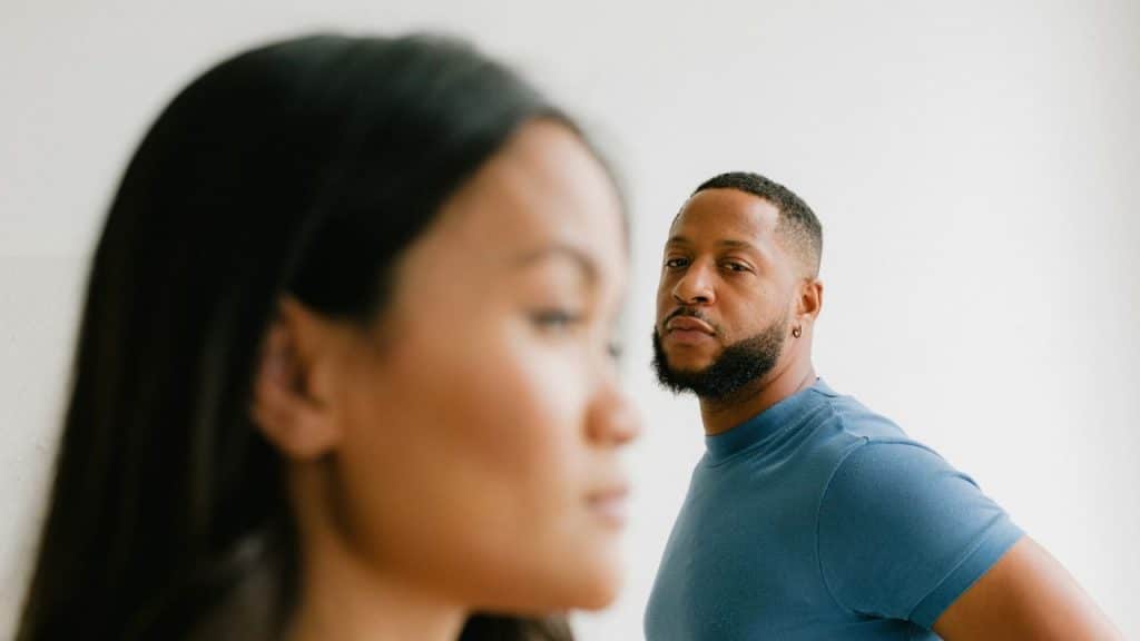 A bearded man in a blue shirt stands in the background, looking directly at the camera, with a woman's head out of focus in the foreground.