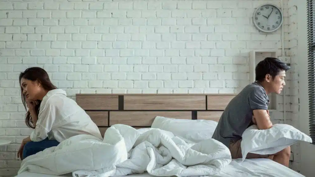 A couple sits on a bed, turned away from each other, looking upset after an argument.