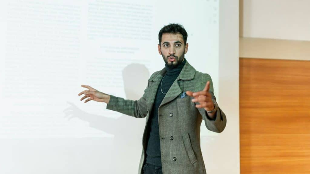 Man in a gray coat and turtleneck presenting in front of a projection screen with text.