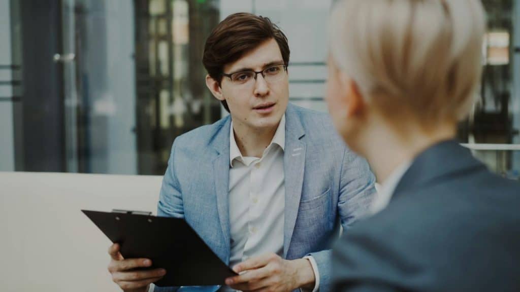 Man in a blue jacket and glasses holding a clipboard while talking to a woman.