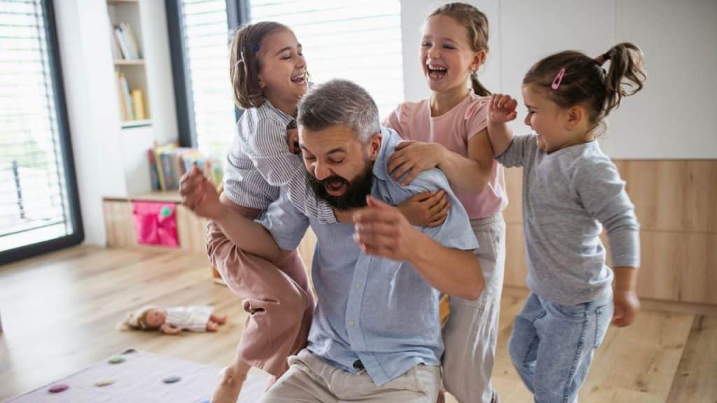 Bearded father laughing while being playfully embraced by three young daughters in a bright room.