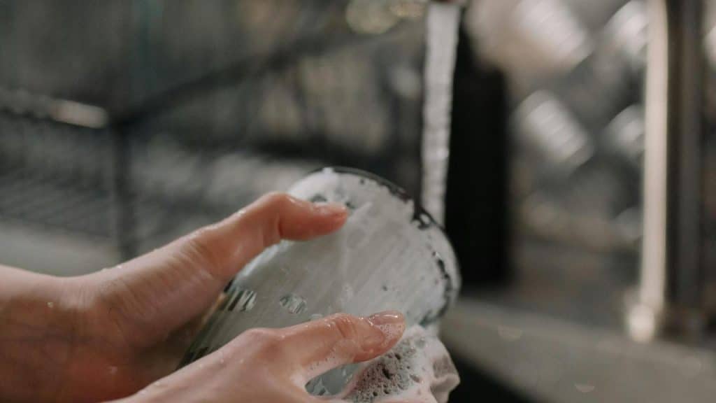 A person washing a soapy glass under running water in a sink.