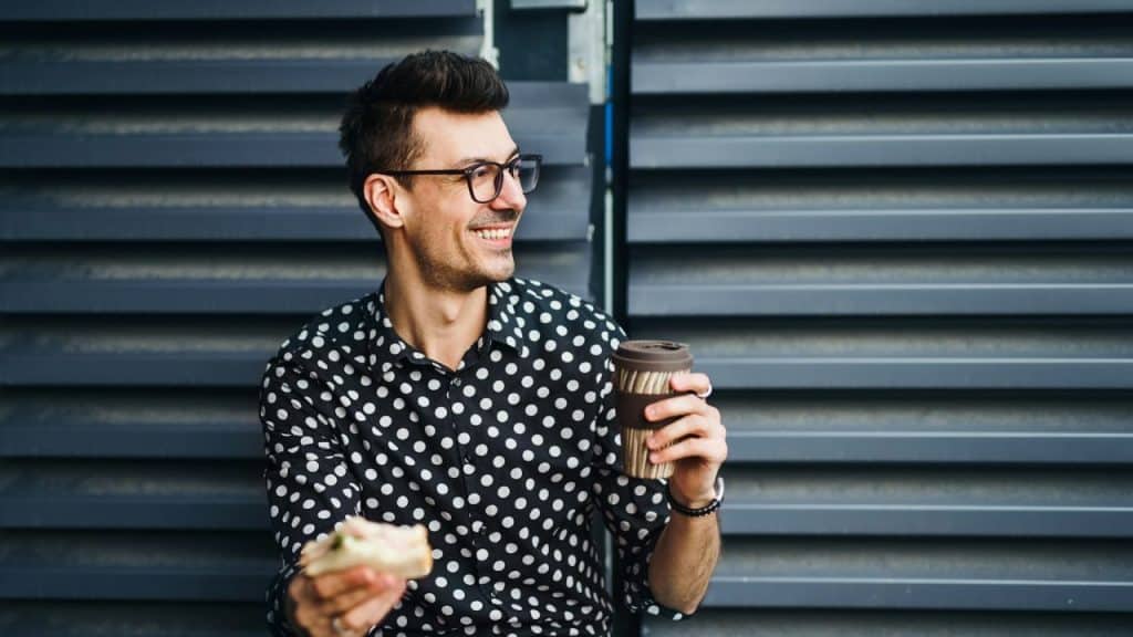 Smiling man in a black and white polka-dot shirt, holding a coffee cup and a sandwich.