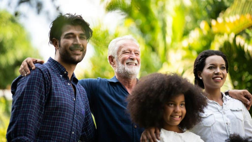 Four smiling people of different ages standing together outdoors in sunlight.