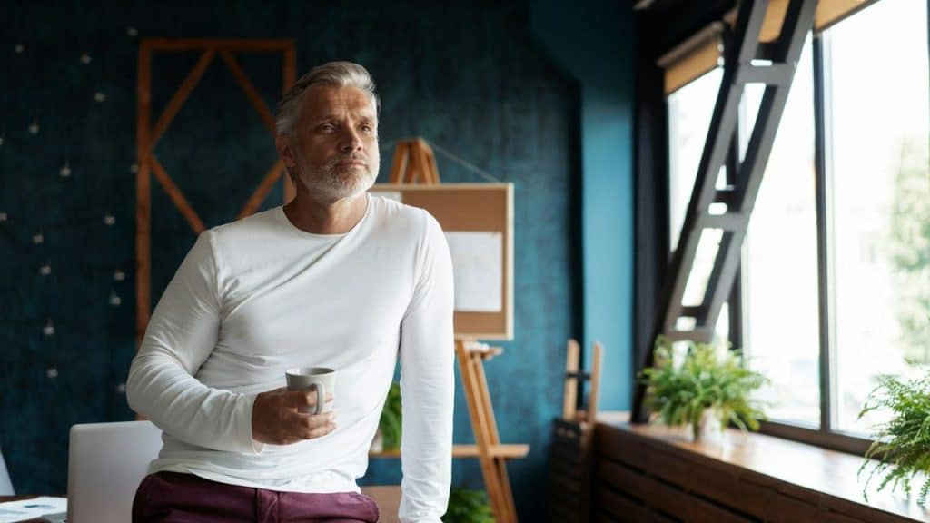 Middle-aged man in a white shirt holding a mug, looking out a large window.