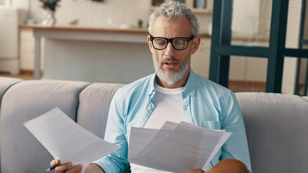 Middle-aged man in glasses holding and reading several papers on a couch.