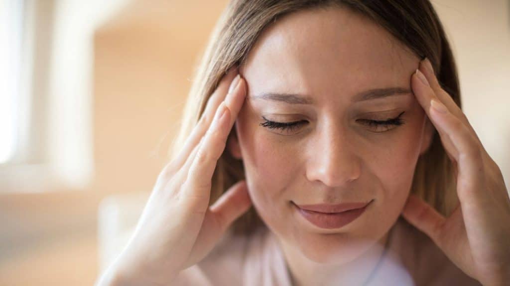 Woman with her eyes closed, pressing her temples with both hands indoors.