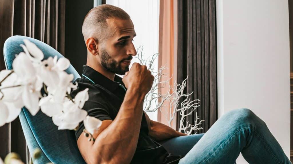 Man with a short haircut and beard sitting in a blue chair, drinking from a cup.