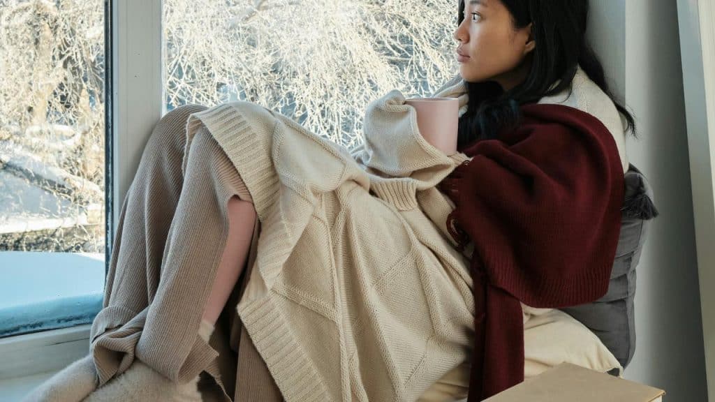 A person wrapped in cozy blankets sits by a window holding a mug and looking outside.