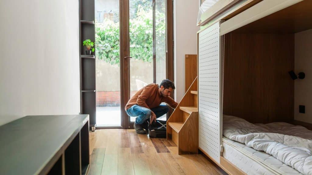 A man crouching in a bright room, fixing or inspecting wooden stairs beside a bed.