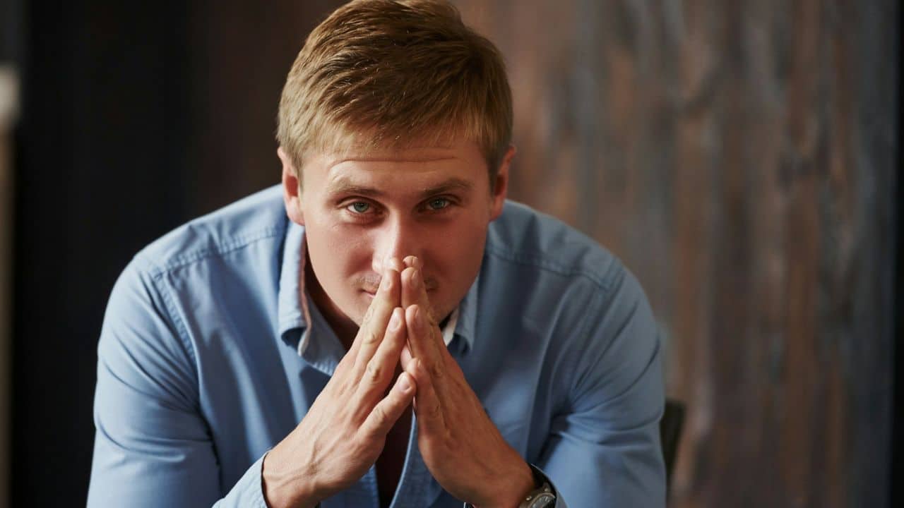 Young man in a blue shirt with hands clasped under his chin looking directly forward.
