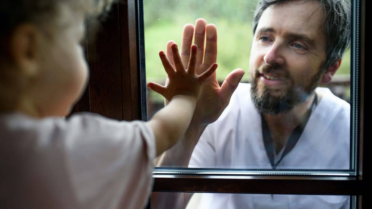 Father and child touching hands on a glass window, smiling at each other.