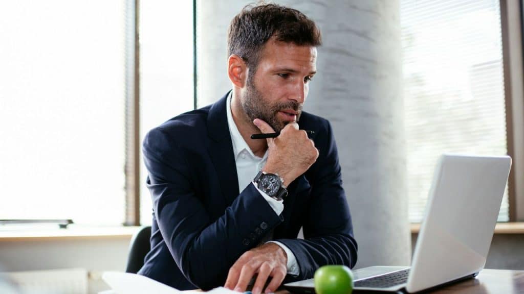 Businessman in a suit and beard working on a laptop at a bright office desk.