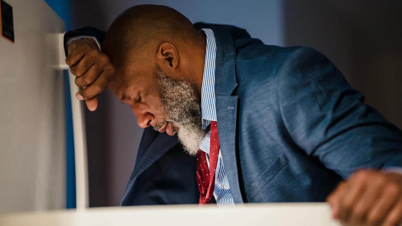 A man in a suit leaning forward with his head down, appearing stressed or exhausted.