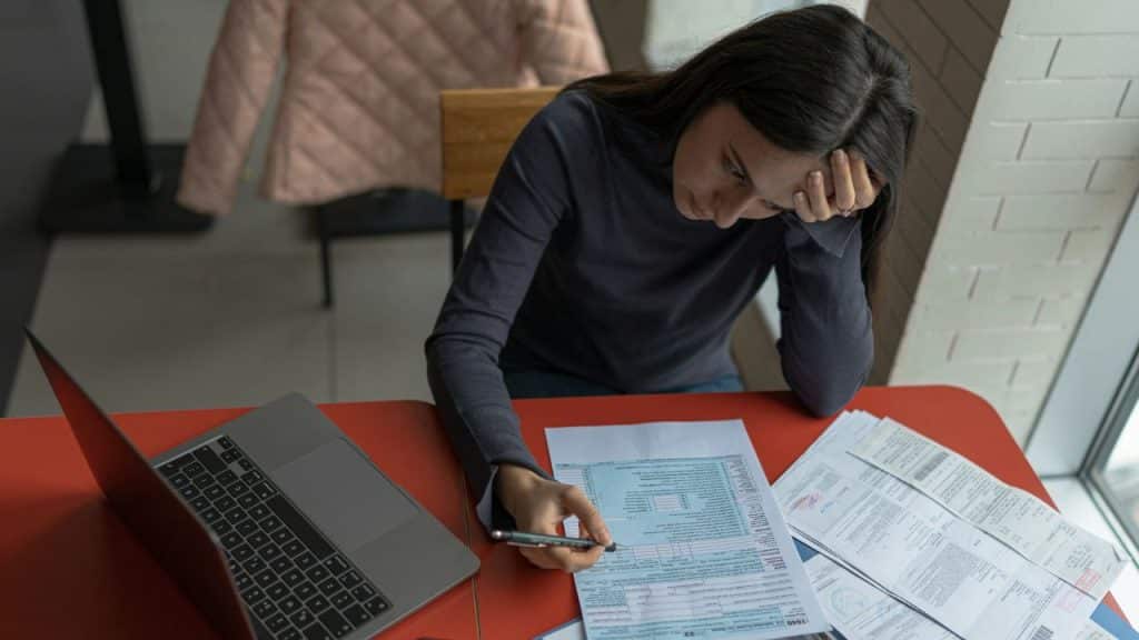 Woman looking stressed while reviewing documents with a pen next to a laptop.