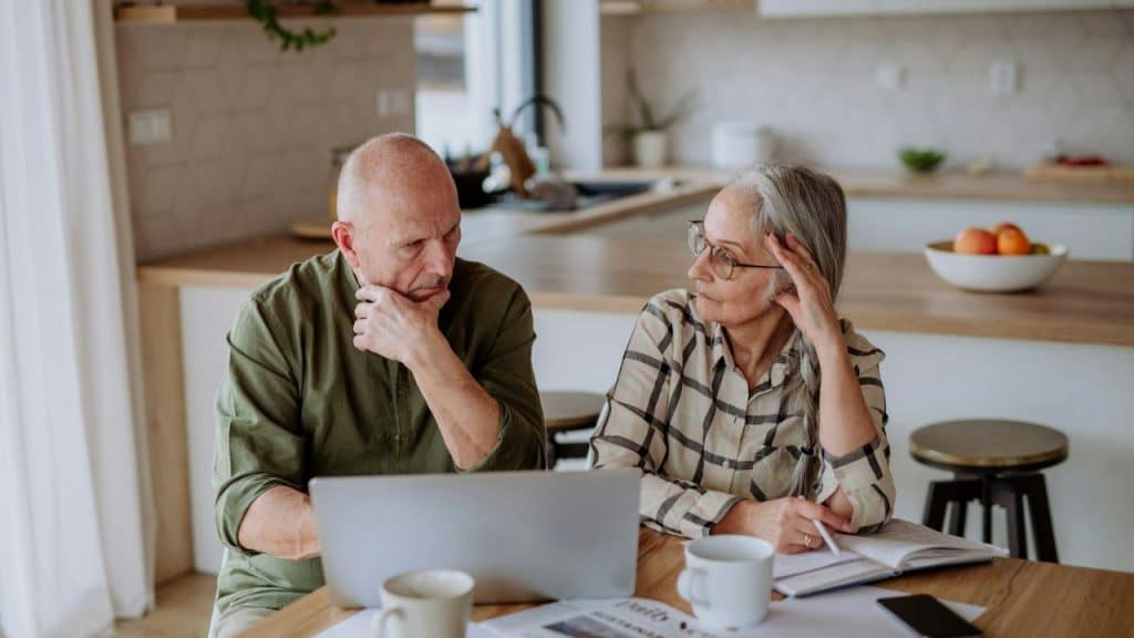 Older couple stressed while working on a laptop at a kitchen counter.