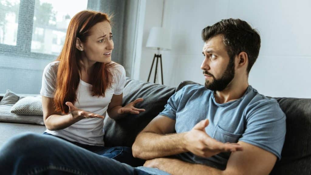 A man and woman sitting on a couch engaged in a tense discussion, gesturing with their hands.