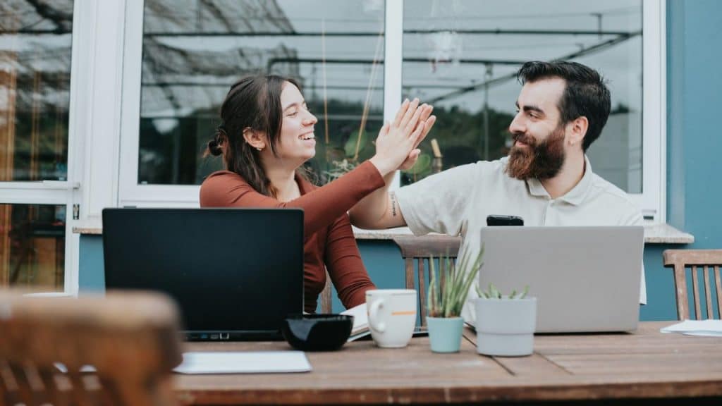 Smiling couple working on laptops outdoors, giving each other a high-five.