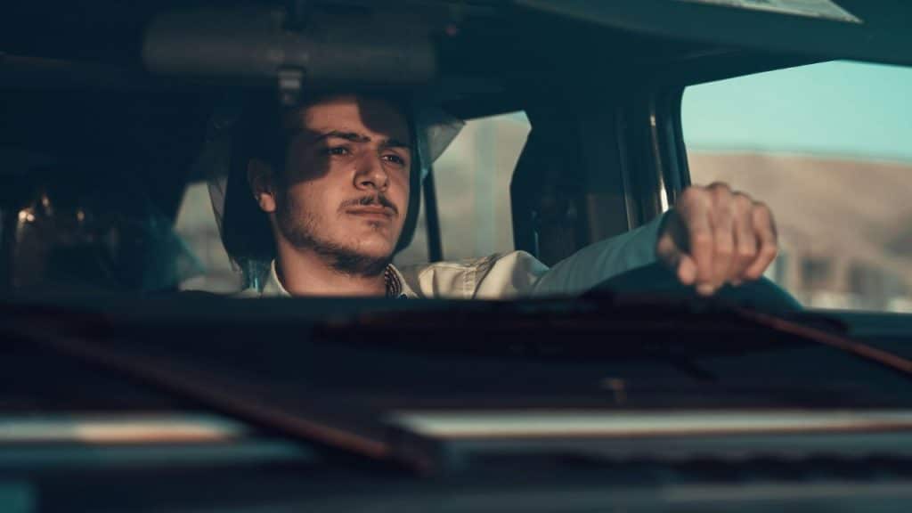 A man with a mustache driving a vehicle, looking ahead with a serious expression.