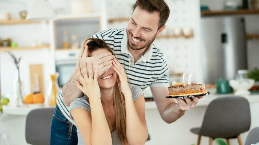 Smiling man covering a woman's eyes while holding a small birthday cake.