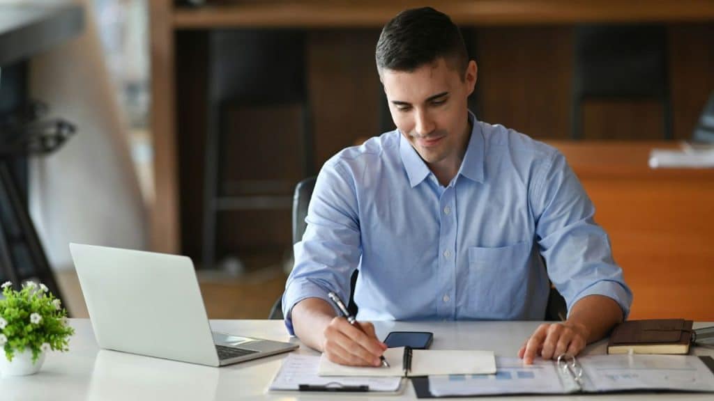 A smiling man in a blue shirt writing in a notebook at a desk with a laptop.