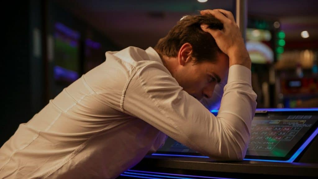 Man in a white shirt with hands on his head, leaning over a glowing electronic screen in a dark room.
