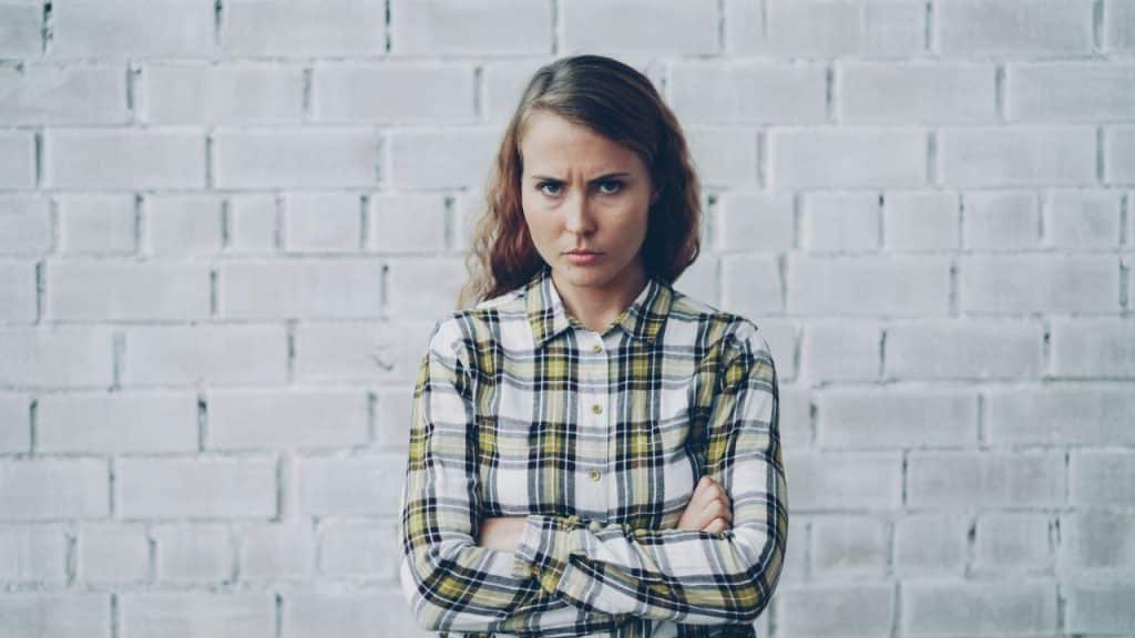 Woman in a plaid shirt with crossed arms, looking angrily at the camera against a white brick wall.
