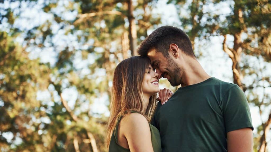 Smiling couple standing close with foreheads touching in a sunny park.