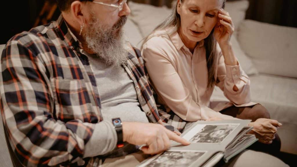 An elderly couple sitting on a couch looking through a photo album together.