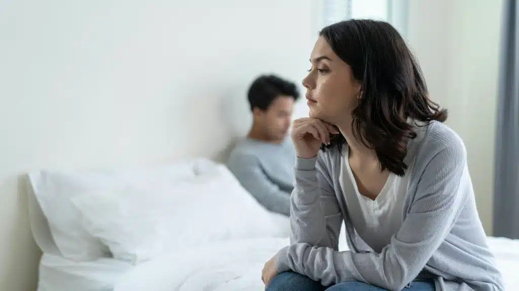 Unhappy woman sitting on the edge of a bed, ignoring the man sitting behind her.