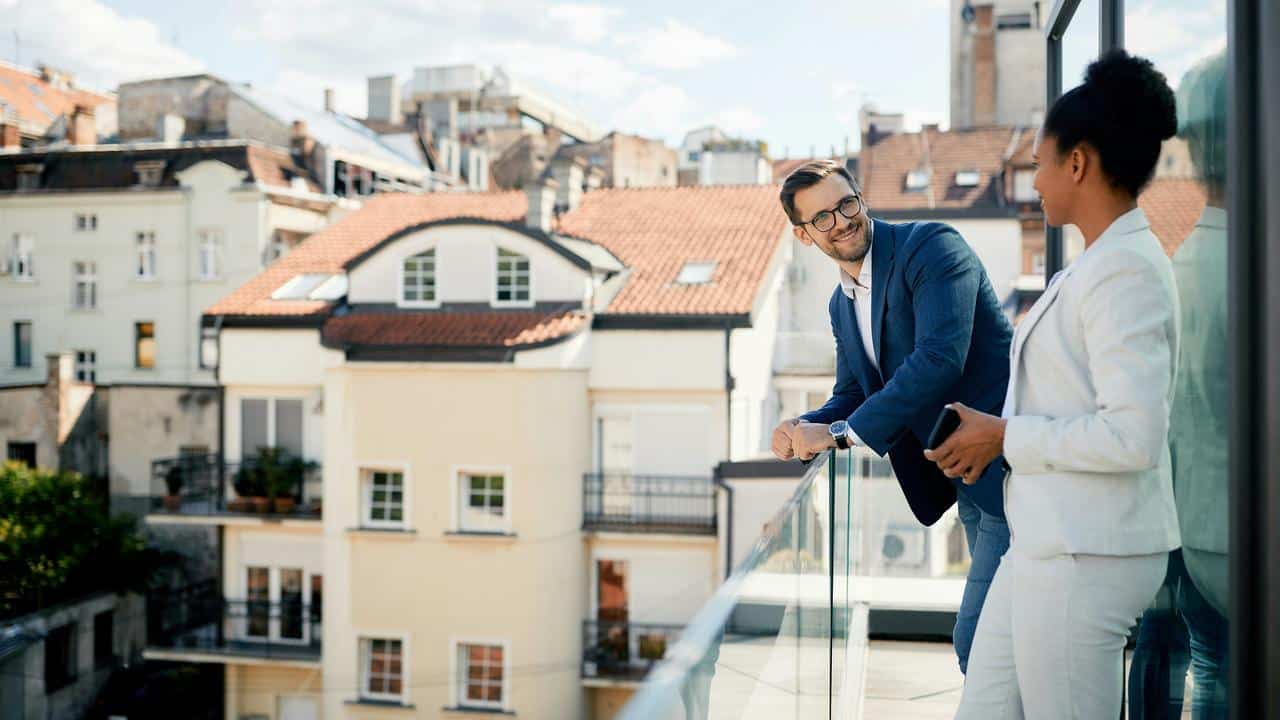 A man and woman in business attire talking on a balcony with city buildings in the background.