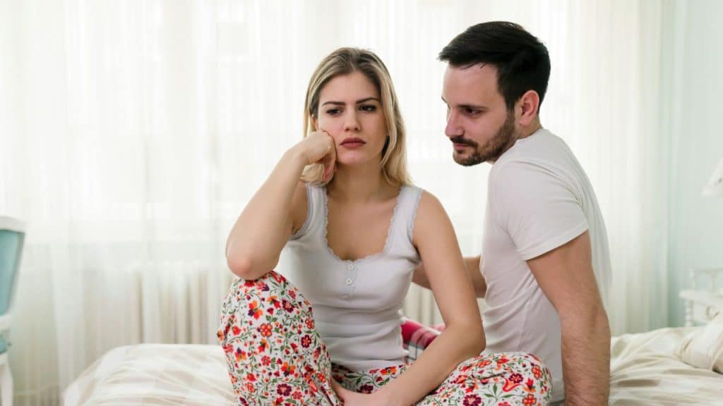 A sad-looking woman sits on a bed while a man looks on with a caring expression.