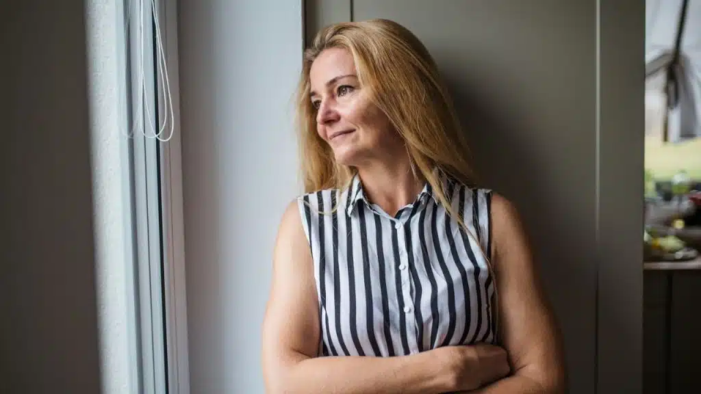 A blonde woman in a black and white striped shirt smiles while looking out a window.