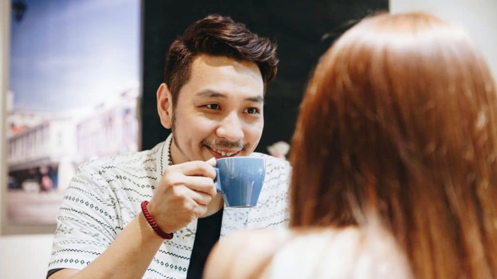 A smiling man holding a blue cup looks at a person with long red hair across a table.