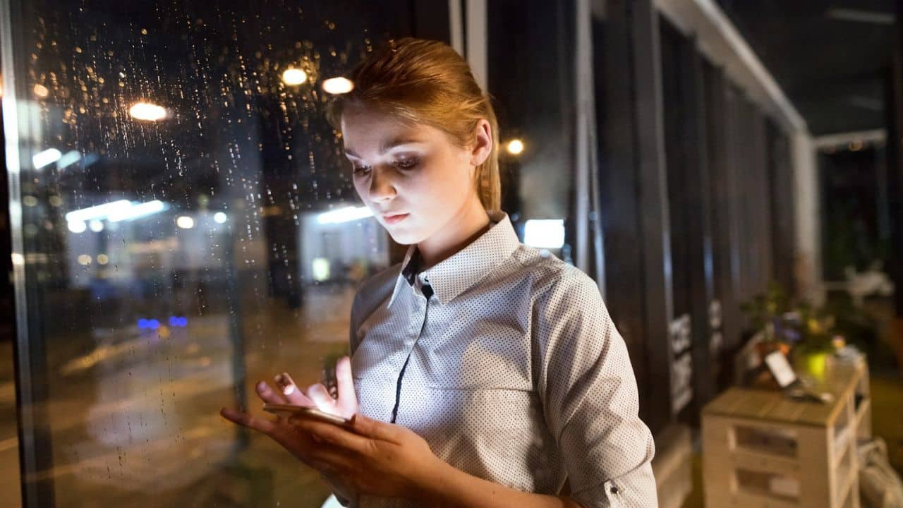 A woman in a white collared shirt looks at a glowing phone screen near a wet window at night.
