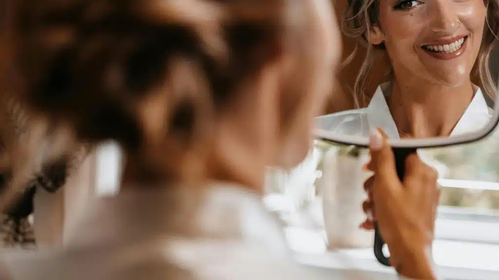 A woman smiling at her reflection while holding a handheld mirror.