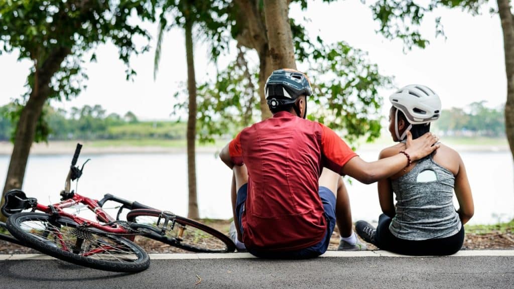 Couple wearing helmets sitting by a path near a lake with a bike resting beside them.