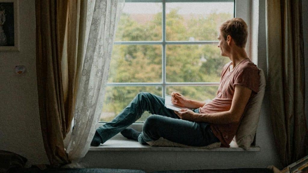 A man sits by a window holding a notebook and looking outside thoughtfully.