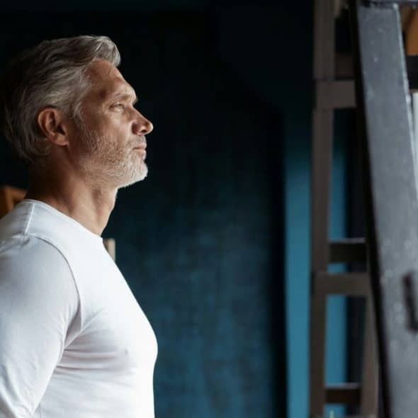 A middle-aged man with gray hair and a white shirt looking out a window.