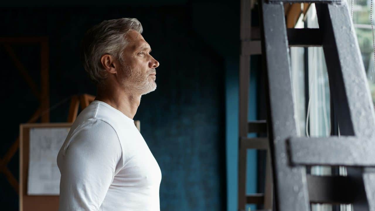 A middle-aged man with gray hair and a white shirt looking out a window.