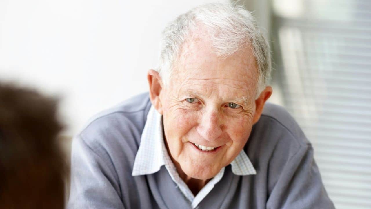 A smiling elderly man with white hair is looking directly at the camera indoors.