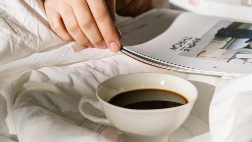 A cup of coffee on a bed beside someone flipping through a magazine.