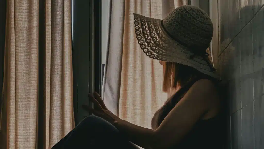 A person wearing a wide-brimmed straw hat is sitting on a windowsill, looking out past closed curtains.