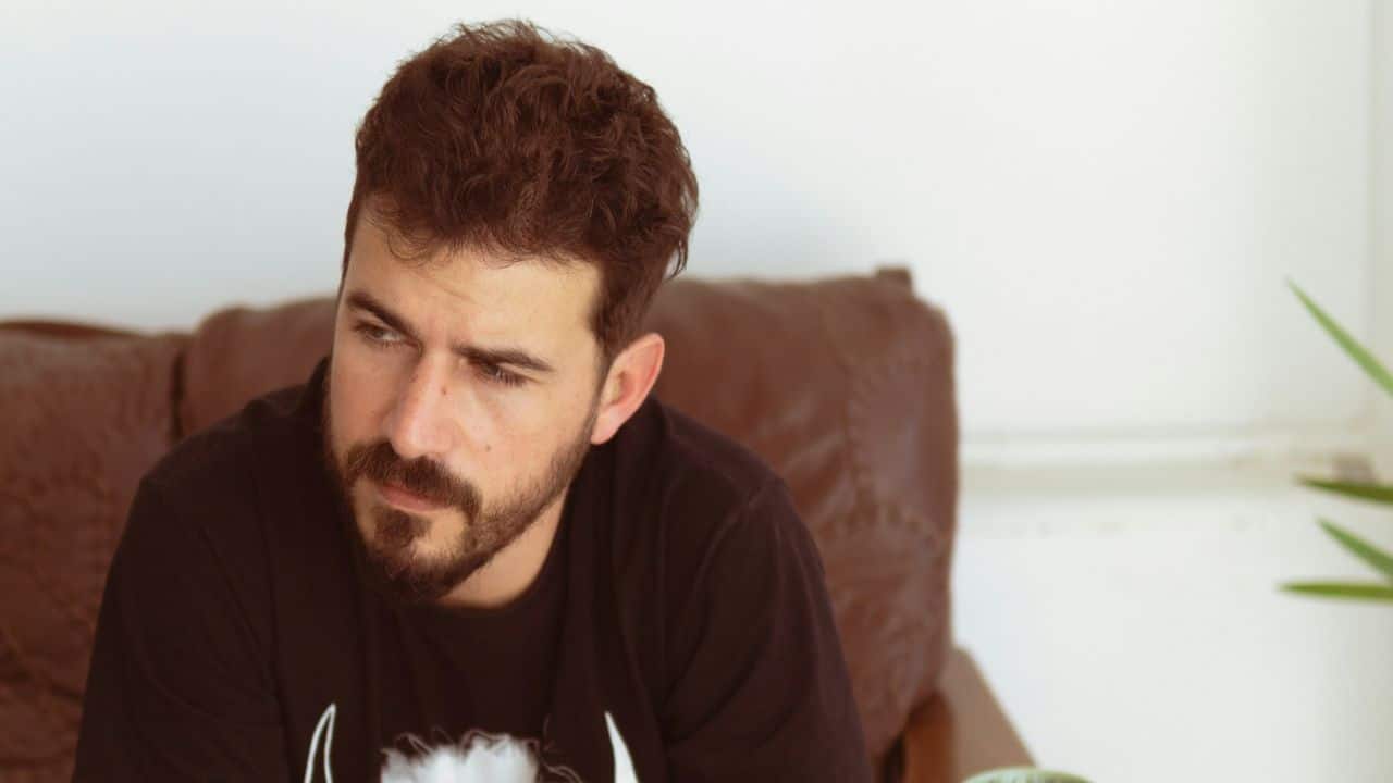 A bearded man with curly hair is sitting, looking thoughtfully to the left.