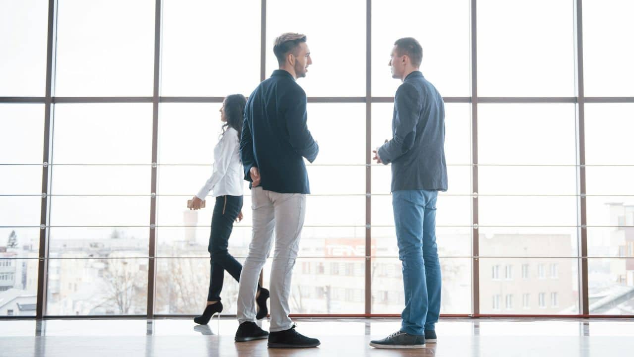 Two men talking in front of a large window while a woman walks past them.