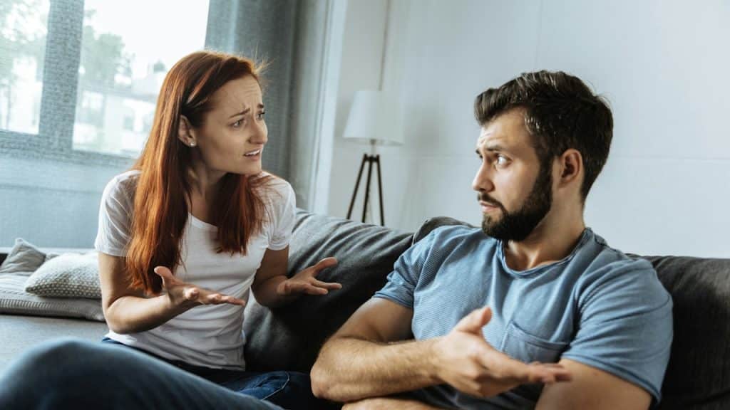 Couple arguing on a couch, both gesturing with their hands during a heated discussion.